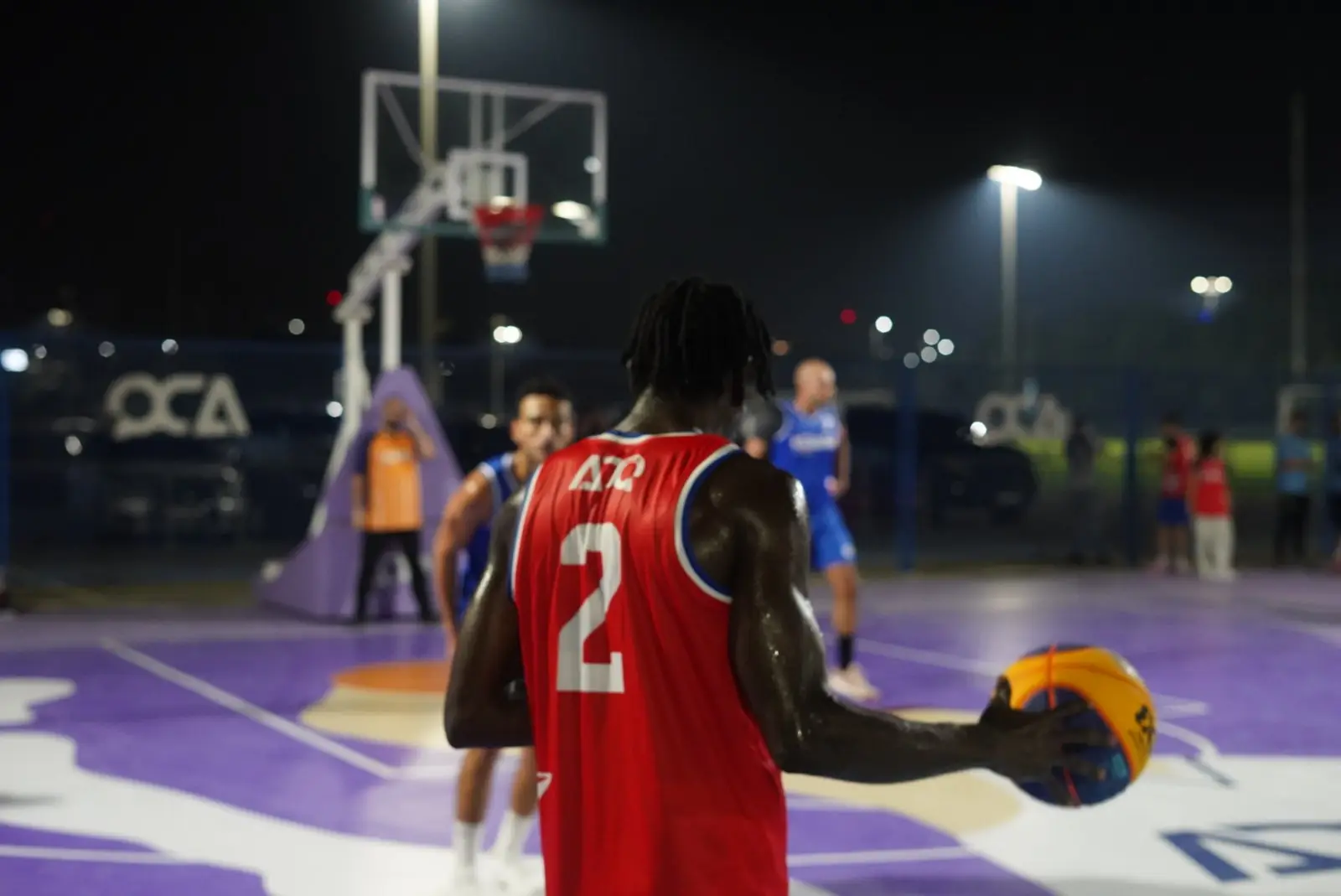 A night basketball game under floodlights, a player in a red number 2 jersey holding the ball